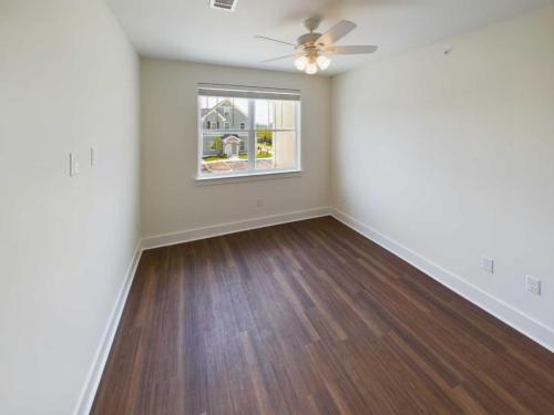 Apartments in Lebanon, IN An empty room with white walls, dark hardwood floors, a ceiling fan with lights, and a window revealing an outside view of houses and greenery. Perfect for those seeking apartments for rent in Lebanon, Indiana.