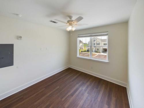 Apartments in Lebanon, IN Empty room with white walls, wood flooring, ceiling fan with lights, and a window overlooking neighboring buildings. The electrical panel is mounted on the wall. Perfect for anyone seeking Apartments For Rent in Lebanon Indiana.