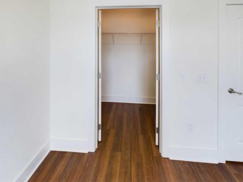 Apartments in Lebanon, IN A room with wooden flooring leads to an open walk-in closet featuring illuminated empty shelves. The walls are painted white, with two light switches visible on the right side of the doorway. Ideal for those looking at apartments for rent in Lebanon, Indiana.