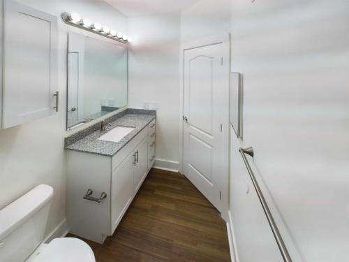 Apartments in Lebanon, IN A modern bathroom in one of the Apartments For Rent in Lebanon, Indiana, features a long countertop and sink, a mirror with lights above, white cabinets, a toilet, and a visible door. The floor is fitted with wood-like tiles for an elegant touch.