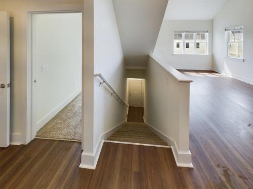 Apartments in Lebanon, IN A home's interior with an open doorway on the left, carpeted staircase leading down the center, and a large room with wooden flooring and sunlight streaming through windows on the right.