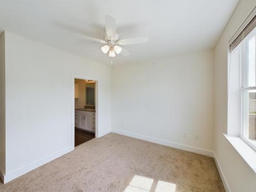 Apartments in Lebanon, IN A vacant bedroom with beige carpet, white walls, and a ceiling fan with lights. An open doorway leads to a bathroom and a window lets in natural light.