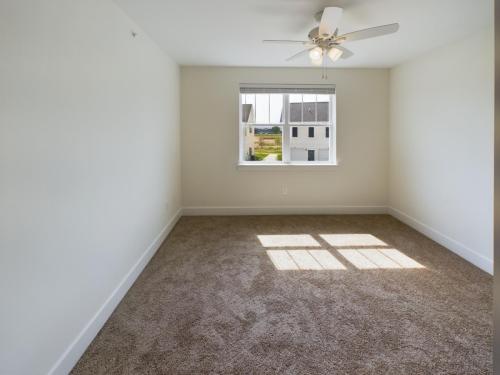 Apartments in Lebanon, IN A sunlit empty room with beige carpet, white walls, ceiling fan, and a window overlooking neighboring houses.