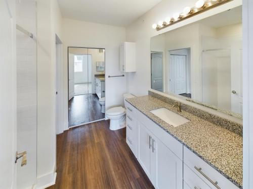 Apartments in Lebanon, IN Modern bathroom featuring white cabinets, a large granite countertop, a wide mirror with vanity lights, a toilet, a shower area, and wood flooring.