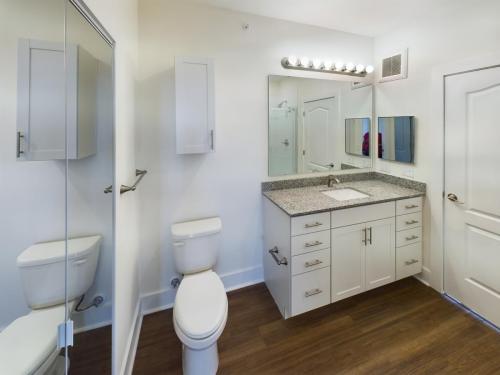 Apartments in Lebanon, IN Modern bathroom with a toilet, sink with granite countertop, cabinets, mirrors, and a shower reflected in a mirrored door. Walls are white and the floor is dark wood.