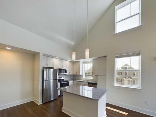 Apartments in Lebanon, IN Modern kitchen with stainless steel appliances, white cabinets, granite countertop island, and hardwood flooring, lit by pendant lights. High ceiling and large windows provide natural light.