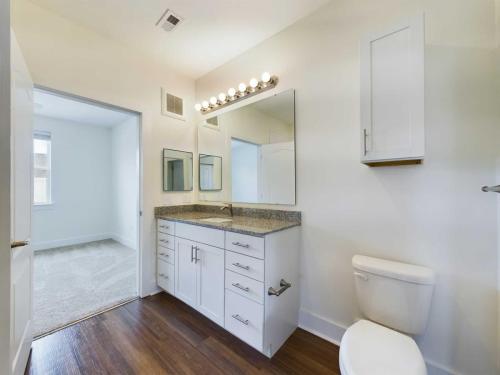 Apartments in Lebanon, IN Spacious bathroom with wooden floor, vanity with granite countertop, multiple mirrors, sink, cabinets, toilet, and doorway leading to an adjacent carpeted room. Bright lighting from vanity bulbs.