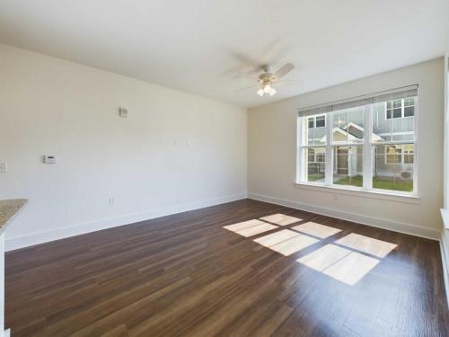 Apartments in Lebanon, IN A vacant, sunlit room with wood flooring, a ceiling fan, large window, and white walls.