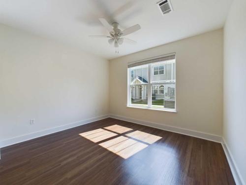 Apartments in Lebanon, IN An empty room with white walls, a brown wood floor, a ceiling fan, and a window letting in natural light. A view of the exterior buildings is visible through the window.