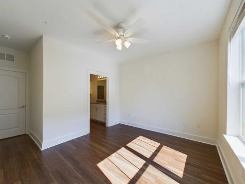 Apartments in Lebanon, IN A bright empty room with hardwood floors, a ceiling fan, a large window, and an open doorway leading to a bathroom with a vanity and lights.