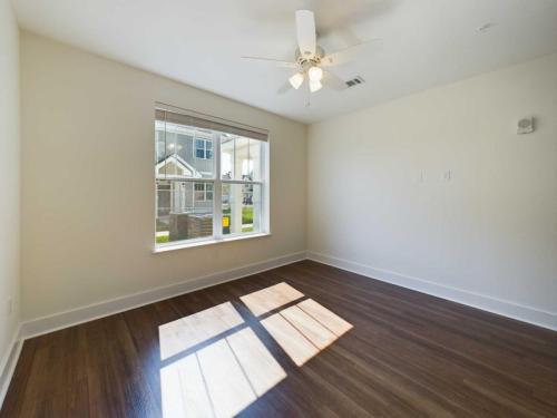 Apartments in Lebanon, IN A clean, empty room featuring a ceiling fan, large window with blinds, hardwood flooring, and white walls, with sunlight streaming in from the window.