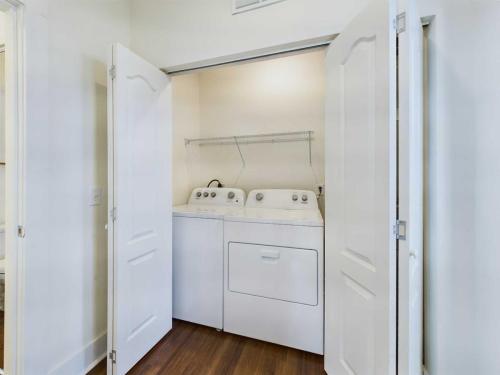 Apartments in Lebanon, IN A laundry closet with open double doors revealing a washing machine and dryer, both white, with a wire shelf above them. The floor is hardwood, and the walls are white.