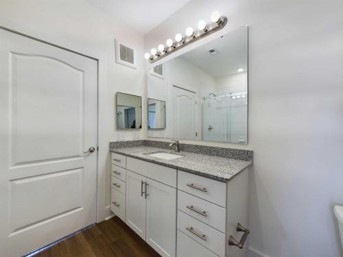 Apartments in Lebanon, IN A modern bathroom features a large mirror with lights above a granite countertop, white cabinets with silver handles, and a door on the left.