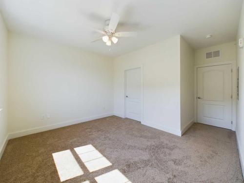 Apartments in Lebanon, IN A clean, empty room with beige carpet, white walls, a ceiling fan with lights, a window, and two white doors.