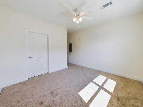 Apartments in Lebanon, IN A vacant room with beige carpet, white walls, a ceiling fan, a closed white door, and a window casting sunlight on the floor.