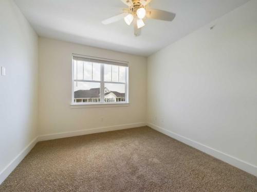 Apartments in Lebanon, IN An empty room with beige carpet, white walls, a ceiling fan with lights, and a window with a view of neighboring buildings.