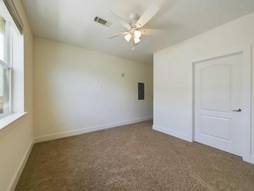 Apartments in Lebanon, IN An empty, carpeted room with white walls, a ceiling fan, a window on the left, and a closed door on the right.