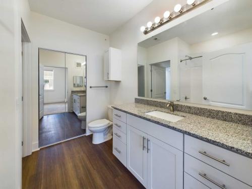 Apartments in Lebanon, IN A modern bathroom featuring a large mirror, white cabinets with granite countertops, a toilet, and a shower with a glass door. The floor is wooden, extending to the adjacent room seen through an open door.