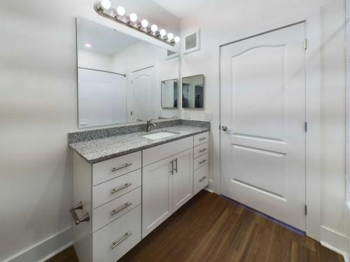 Apartments in Lebanon, IN A modern bathroom featuring a white vanity with a granite countertop and chrome faucet, a large mirror with a row of lights above, wooden flooring, and a white door on the right.