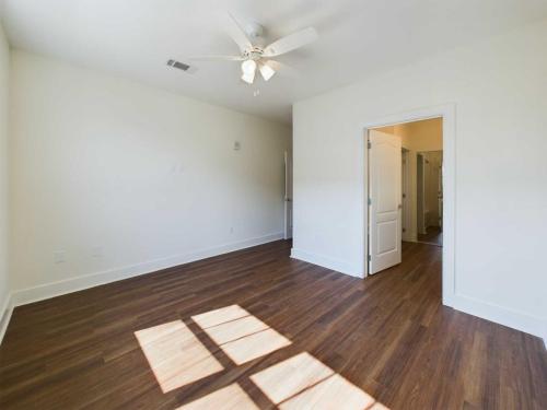 Apartments in Lebanon, IN Empty room with white walls, a ceiling fan, and brown wood-like flooring. Sunlight through a window creates patterns on the floor. An open door reveals part of a connected bathroom.