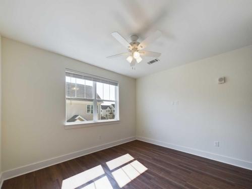 Apartments in Lebanon, IN A bright, empty room with white walls, a ceiling fan, a window with blinds, and wooden flooring. Sunlight pours through the window creating a shadow on the floor.