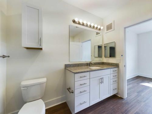 Apartments in Lebanon, IN Modern bathroom with a white toilet, a granite countertop vanity with drawers, a large mirror with lights above it, and a wooden floor. An adjacent room is visible through an open door.