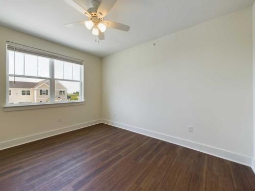 Apartments in Lebanon, IN An empty room with white walls, a ceiling fan with lights, and a large window allowing natural light. The floor is covered with dark wood flooring.