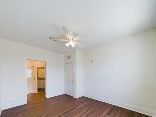 Apartments in Lebanon, IN A white-walled, empty room with brown wood flooring, a ceiling fan, and two doors; one open showing a glimpse of a bathroom and the other closed.