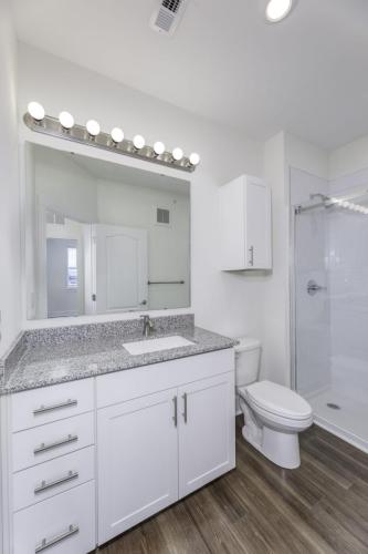 Apartments in Lebanon, IN Modern bathroom with white vanity cabinets, granite countertop, large mirror with light bulbs, toilet, and glass-enclosed shower. Wood-like flooring and white walls complete the clean design.