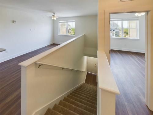 Apartments in Lebanon, IN A view of an empty, well-lit room with wooden flooring. There is a staircase with carpeting leading down and windows with white trim. A ceiling fan is visible on the left side of the room, making it an ideal option among apartments for rent in Lebanon Indiana.